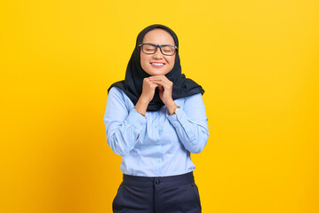 Portrait of happy young Asian woman keeps hands under chin, looks aside, remembers pleasant moment isolated on yellow background