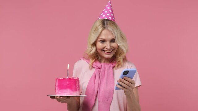 Excited elderly blonde woman lady 40s years old wears t-shirt birthday hat hold cake with candle use mobile cell phone read sms chat text isolated on plain pastel light pink background studio portrait