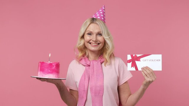 Elderly blonde woman lady 40s years old in t-shirt birthday hat look at cake with candle gift certificate coupon voucher card for store isolated on plain pastel light pink background studio portrait