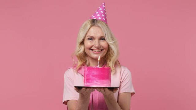 Charming fun excited elderly blonde woman lady 40s years old wears t-shirt birthday hat look camera hold cake with candle wink blink eye isolated on plain pastel light pink background studio portrait