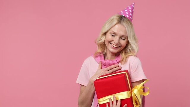 Side view profile elderly blonde woman lady 40s years old in t-shirt birthday hat catch red present box with gift ribbon bow say thank you isolated plain pastel light pink background studio portrait