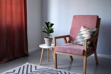 Comfy wooden armchair with houseplant on table near light wall