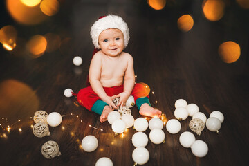 Fototapeta na wymiar A small child, newborn, baby or toddler in a Santa Claus hat is sitting on the floor at home and playing with Christmas toys, there is a garland nearby, Christmas lights around, bokeh.