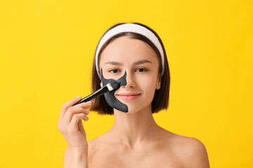 Young woman applying activated charcoal mask on her face against color background