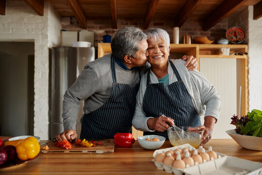Biracial elderly couple happily cooking in the kitchen. Husband kissing wife on the cheek, healthy retired lifestyle. - Powered by Adobe