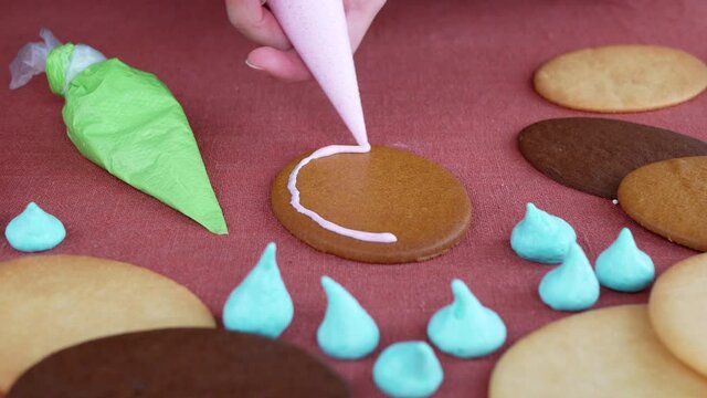 Person using pink flood consistency icing to pipe a border around the edges of the cookie, then filling in it. Hands outline and fill gingerbread with royal icing, close up