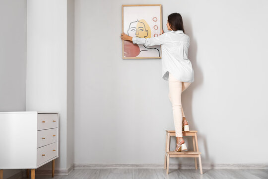 Woman with stepladder hanging painting on light wall