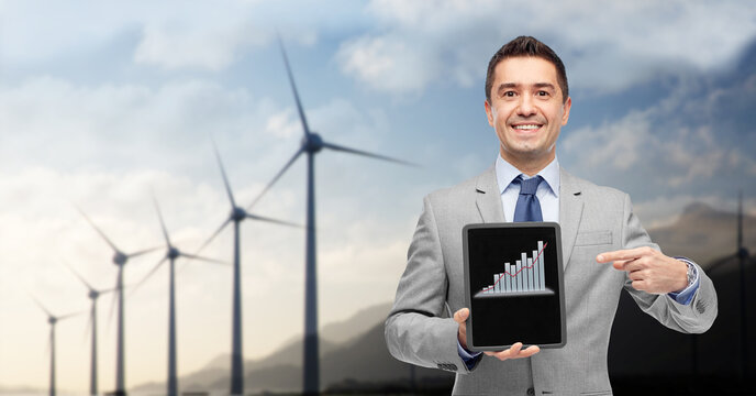business, technology and green energy concept - smiling businessman with charts on tablet pc computer screen over wind turbines on background