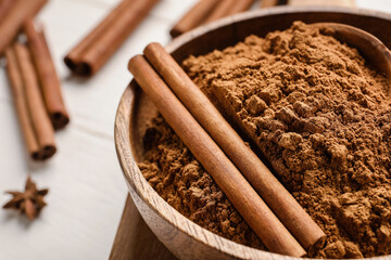 Bowl with cinnamon powder and sticks on white wooden background, closeup