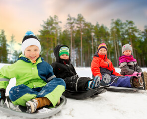 childhood, sledging and season concept - group of happy little kids sliding on sleds in winter over snowy forest or park background