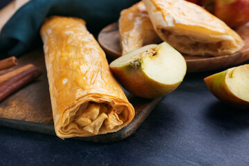Wooden board with tasty apple strudel and fresh fruits on black background, closeup