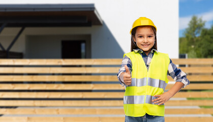 building, construction and profession concept - smiling little girl in protective helmet and safety vest showing thumbs up over living house background