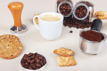 Cup of espresso, crackers, cookies, holder with ground coffee, tamper and coffee beans on table.