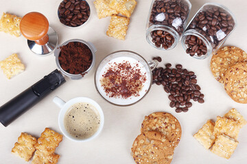 Cup of coffee, cappuccino, crackers, cookies, holder with ground coffee, tamper and coffee beans on table. View from above.