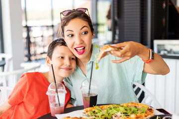 Happy cheerful mommy and her child eat delicious Italian pizza in a pizzeria or cafe. Joint leisure and fast food concept - obrazy, fototapety, plakaty
