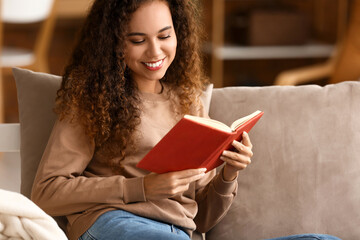 Naklejka premium Smiling young African-American woman reading book on sofa at home