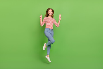 Fototapeta na wymiar Photo of cheerful friendly lady jump show v-signs wear striped shirt jeans sneakers isolated green color background