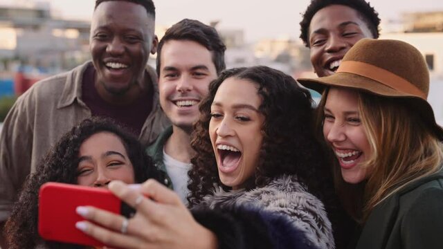 Diverse group of adult friends taking a selfies on a cellphone, laughing on a rooftop at a party at dusk