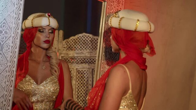 a blonde in a turban and a shiny dress dances with a red scarf in front of a mirror