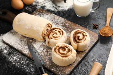 Wooden board with uncooked cinnamon rolls on black background