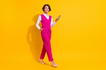 Photo of charming shiny young lady wear pink suit smiling walking typing modern device empty space isolated yellow color background