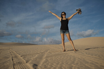 Fototapeta na wymiar Woman in the free pose, rising hands up, stands on the dune. Happy girl on vacation wearing shorts on the background of the sandy desert and sky.