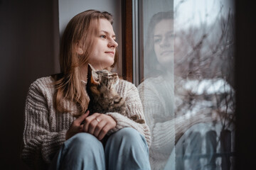 Young cheerful girl sitting at home on the windowsill in a warm sweater playing with a gray cat on a winter day