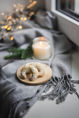Weihnachtliches Stilleben mit einer brennenden Kerze und Vanillekipferl auf einem Teller auf einer Fensterbank mit einer grauen Wolldecke.