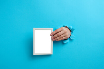 female hand holding empty white wooden frame, body part sticking out of torn hole in blue paper background