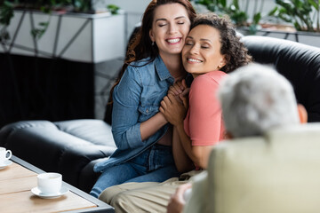 happy multiethnic lesbian women with closed eyes holding hands while sitting on couch near blurred psychologist