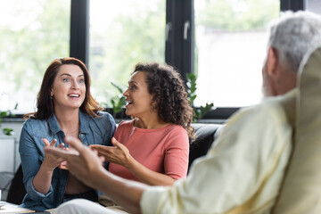excited multiethnic lesbian couple gesturing while talking with blurred psychologist during appointment