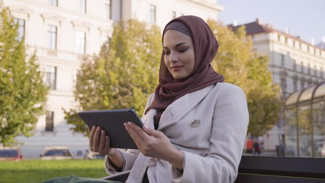A young beautiful Muslim woman looks at a tablet with a smile as she sits in a park in an urban area - closeup from below