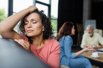 depressed african american woman sitting with closed eyes near girlfriend and psychologist on blurred background