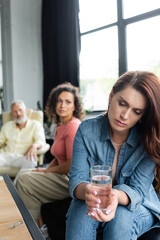 frustrated woman holding glass of water near african american girlfriend and psychologist on blurred background