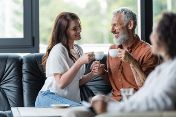 cheerful couple with coffee cups holding hands near blurred african american psychologist