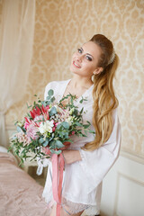Pretty young Bride. Blonde-haired woman with wedding hair-style with a long tail. Boudoir morning of the bride. Looking on her bouquet