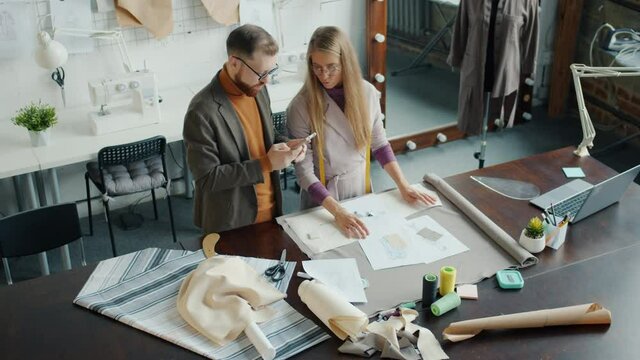 Creative dressmakers girl and guy are discussing new collection choosing material and using smartphone at work. People and tailoring concept.