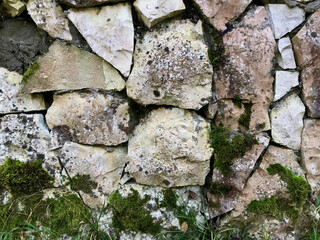 The stone fence covered with green moss. Abstract textured background of stone wall. Old medieval stone wall along a street. Photo closeup of aged building made of stone masonry  outside on cityscape 