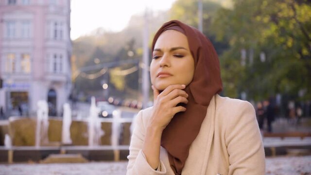 A young beautiful Muslim woman thinks about something in a street in an urban area - a fountain in the blurry background - closeup
