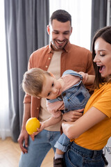 Excited woman holding toddler kid with lemon near blurred husband at home