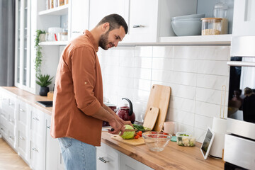 Side view of smiling man looking at digital tablet while cutting vegetables in kitchen