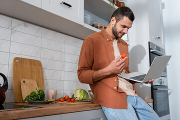 Young man holding cherry tomato and laptop near vegetables in kitchen