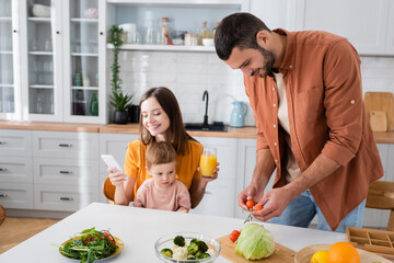 Young man holding cherry tomatoes near smiling wife using smartphone and son at home