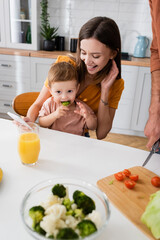 Happy mother holding smartphone and looking at child eating broccoli at home