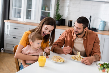 Smiling woman feeding pasta to son near husband and orange juice in kitchen