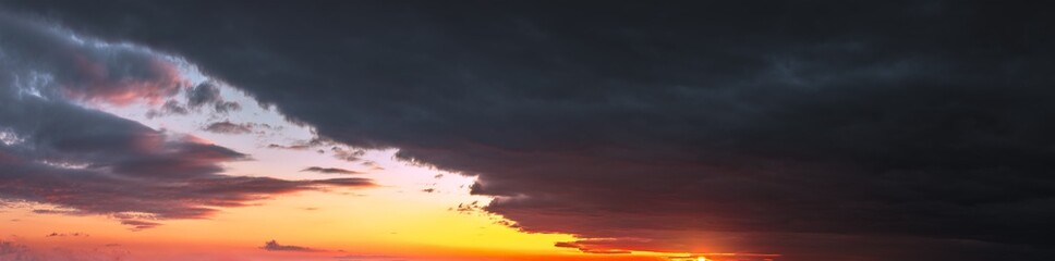 Panorama of dramatic colorful sunset with dark and bright clouds. The sun's rays are breaking through the cloudy sky. Dark clouds against a bright saturated sky.