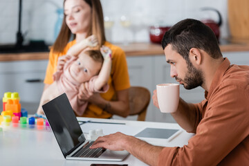 Freelancer holding cup and using laptop near family and building blocks at home