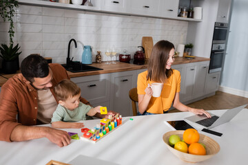 Freelancer with cup using laptop while husband and son playing game in kitchen