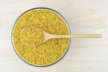 Bulgur groats in a glass round bowl with a wooden spoon on a light wooden background. Dry groats made from boiled, dried and crushed wheat. Healthy carbohydrates. Top view