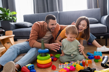 Positive parents hugging toddler kid near toys and bike in living room